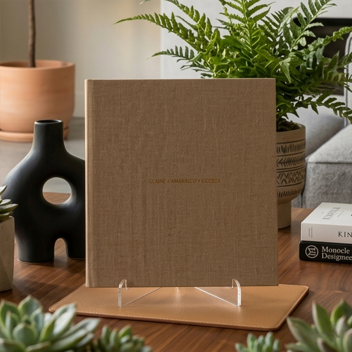 Fotolivro de arte com capa de tecido, exibido em mesa de madeira com plantas decorativas ao fundo.